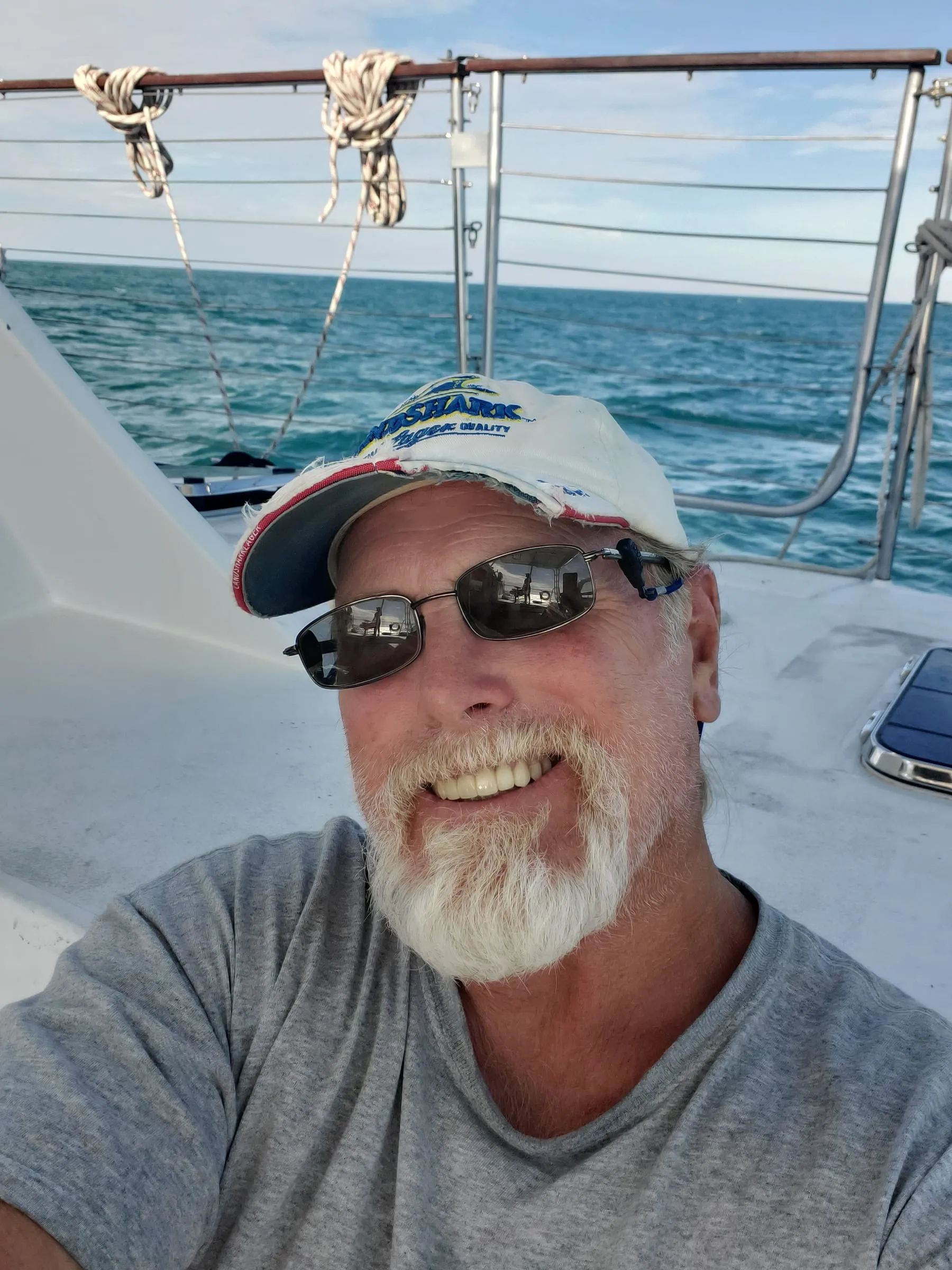 Captain James Lowe, white-bearded in sunglasses and a well-worn cap, smiling aboard a sailboat