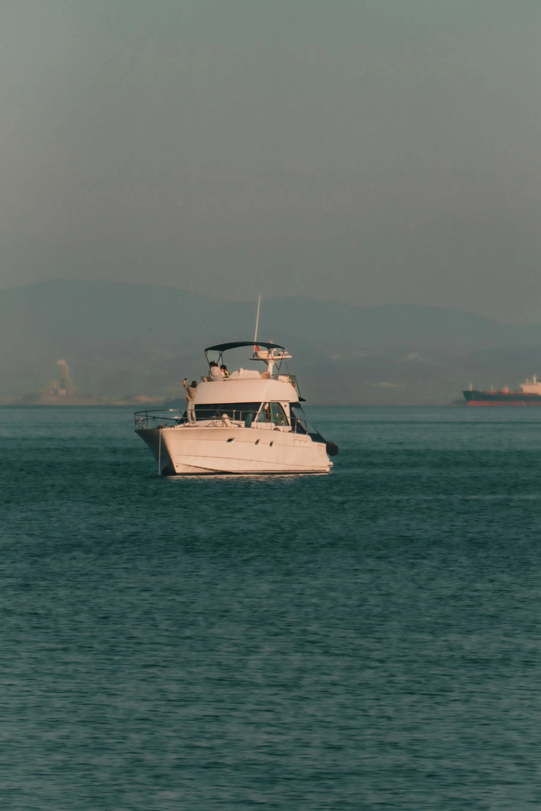 A flybridge motor yacht underway in warm haze, with a ship on the horizon