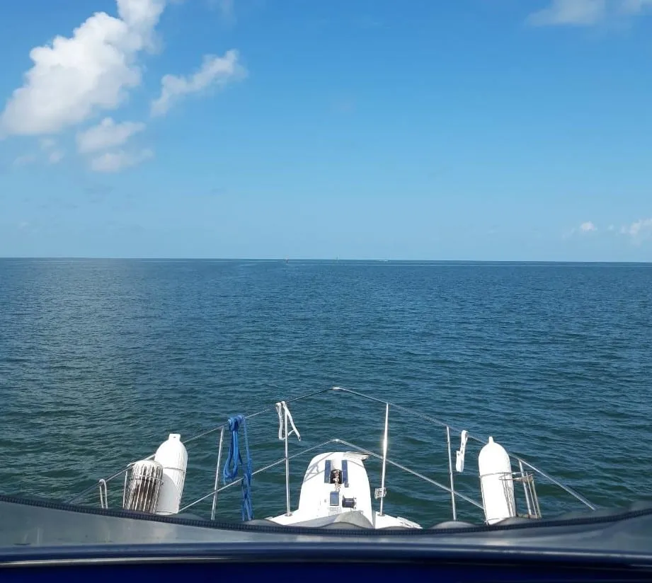 The view forward over a boat’s bow rail and dinghy toward open water and distant channel markers